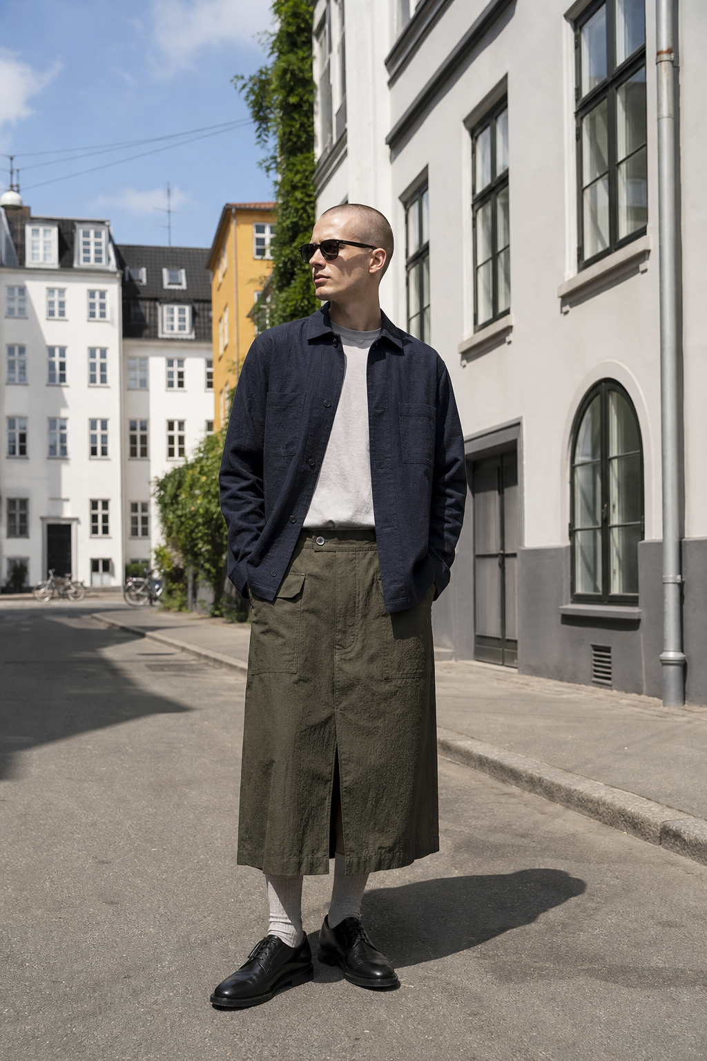 Editorial-Foto eines Mannes in Kopenhagen mit olivfarbenem langem Micro-Seersucker-Rock, navy Overshirt, grauen Kniestruempfen und schwarzen Derby-Boots.